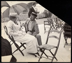 Homme et femme assis dehors sur des chaises pliantes avec une femme tenant un parasol, Narragansett, Rhode Island, 1891